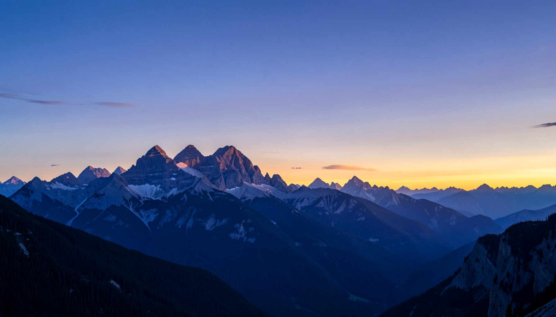 Panoramic banner of Banff's mountain peaks at dusk, layered in blues and purples with a soft golden horizon