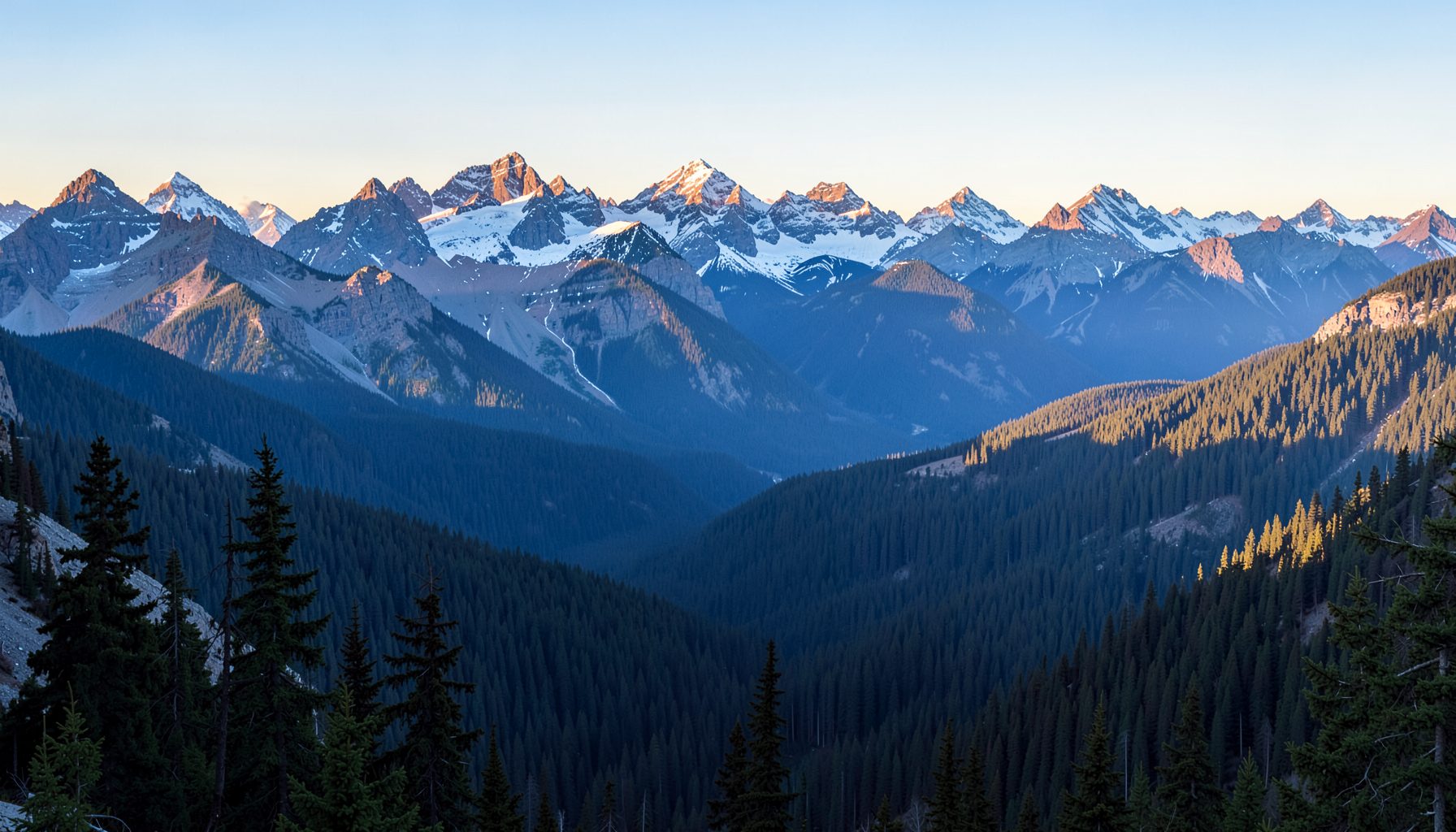 Panoramic banner of the Canadian Rockies at sunrise, layers of pine forest and snow-dusted peaks in soft blue and gold light